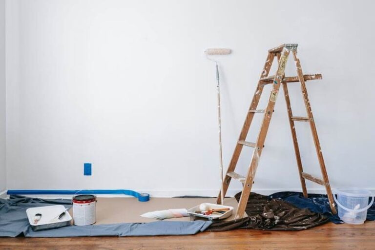 Ladder and rooler standing against wall. Paint canm, floor covering, and brush sitting on florr getting ready to paint.